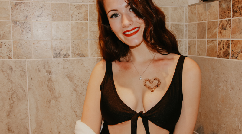 A woman stands in her shower wearing a black bikini top. She's smiling at the camera. There is a heart shape of Koffee Beauty coffee scrub on her chest. To the left of the photo is the title of the article 